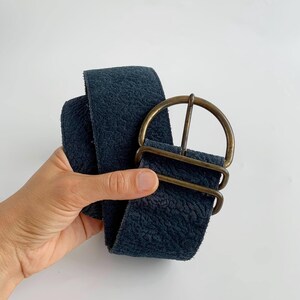 May include: A wide, navy blue belt with a textured surface and a large, antique brass buckle. The belt is held up against a white background, showcasing its colour and design. The buckle has a rounded shape.