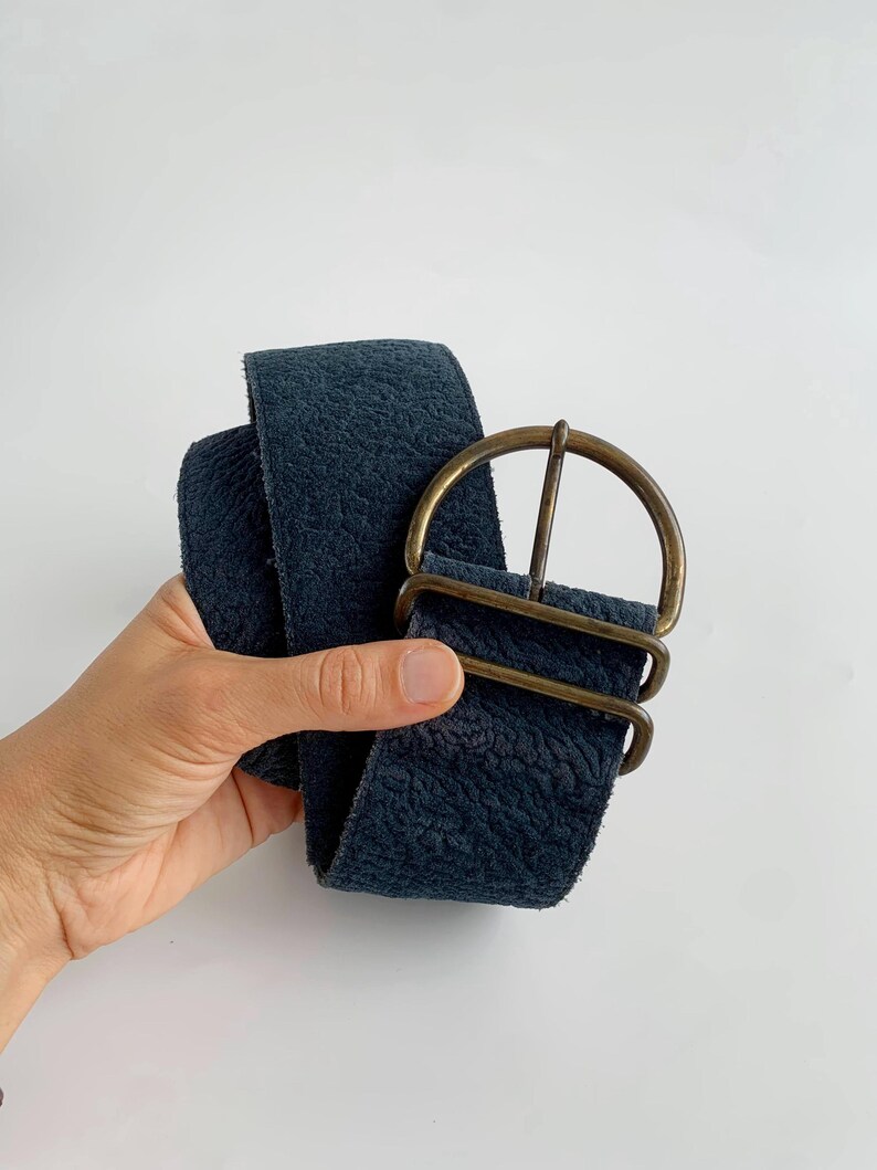 May include: A wide, navy blue belt with a textured surface and a large, antique brass buckle. The belt is held up against a white background, showcasing its colour and design. The buckle has a rounded shape.