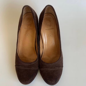 May include: A pair of brown suede heeled shoes with a rounded toe. The shoes have a brown leather insole with the brand name "Salamander" printed on them.