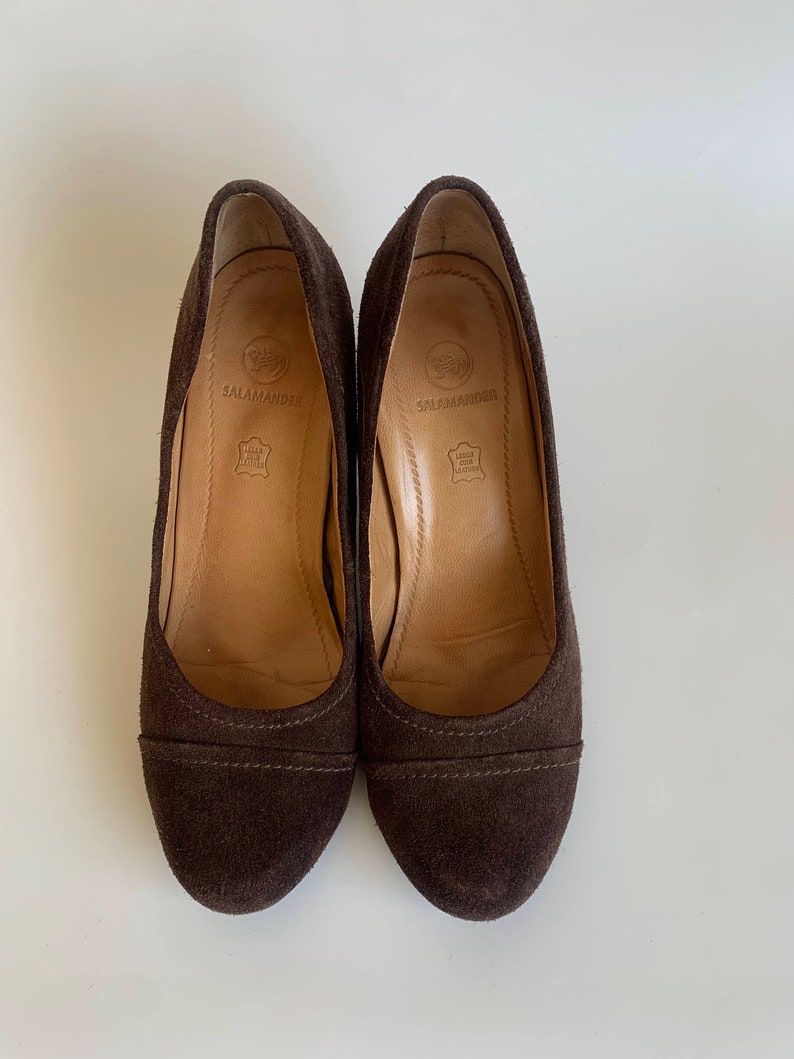 May include: A pair of brown suede heeled shoes with a rounded toe. The shoes have a brown leather insole with the brand name "Salamander" printed on them.