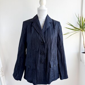 Pode incluir: Um blazer azul marinho com um design texturizado e canelado verticalmente. O blazer tem uma gola entalhada, três botões e dois bolsos frontais. A peça de roupa é exibida em um manequim branco.
