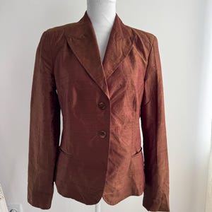 Könnte beinhalten: Ein brauner Blazer mit Zwei-Knopf-Verschluss und zwei Vordertaschen. Der Blazer wird auf einer weißen Schaufensterpuppe präsentiert. Der Stoff hat einen subtilen Glanz, der auf Seide hindeutet. Der Blazer hat ein klassisches Revers-Design.