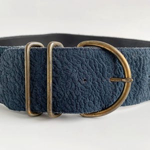 May include: A wide, textured navy blue belt with a brass-coloured buckle and two matching loops. The belt's buckle is a D-shape, and the material has a slightly worn appearance. The belt is designed to be worn around the waist.