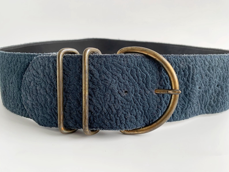 May include: A wide, textured navy blue belt with a brass-coloured buckle and two matching loops. The belt's buckle is a D-shape, and the material has a slightly worn appearance. The belt is designed to be worn around the waist.