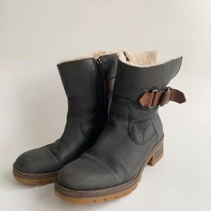 May include: A pair of black leather mid-calf boots featuring a side zip, a brown leather strap with a metal buckle detail at the ankle, and a thick, textured gum-coloured rubber sole. The interior is lined with a light-coloured fleece material.