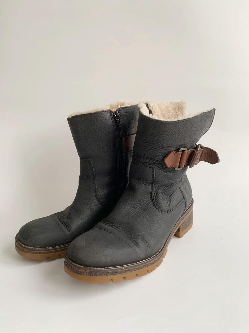 May include: A pair of black leather mid-calf boots featuring a side zip, a brown leather strap with a metal buckle detail at the ankle, and a thick, textured gum-coloured rubber sole. The interior is lined with a light-coloured fleece material.