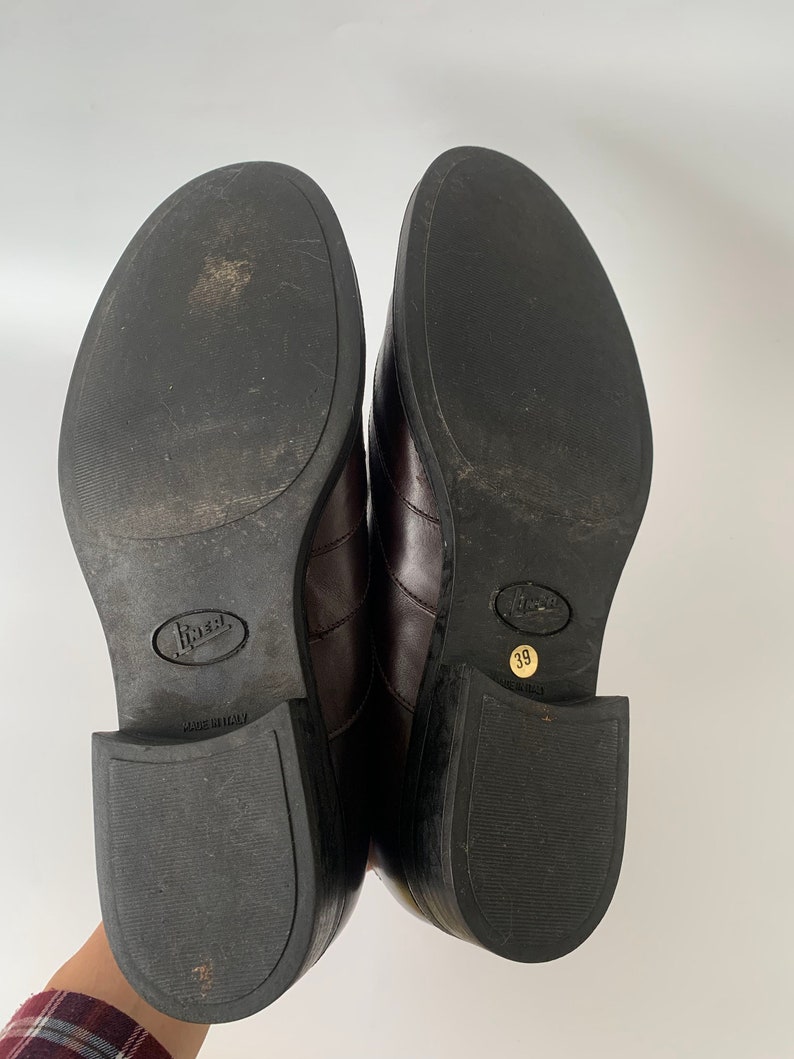 K&ouml;nnte beinhalten: Ein Paar braune Lederschuhe mit schwarzer Gummisohle. Die Schuhe haben eine spitze Zehenpartie und einen niedrigen Absatz. Die Sohlen sind mit "Made in Italy" und "39" gekennzeichnet.