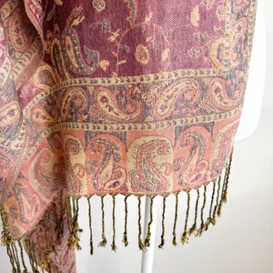 May include: A close-up of a paisley patterned scarf in shades of pink, peach, and purple. The scarf has a fringed edge with gold-colored tassels. The fabric has a woven texture and a decorative border.