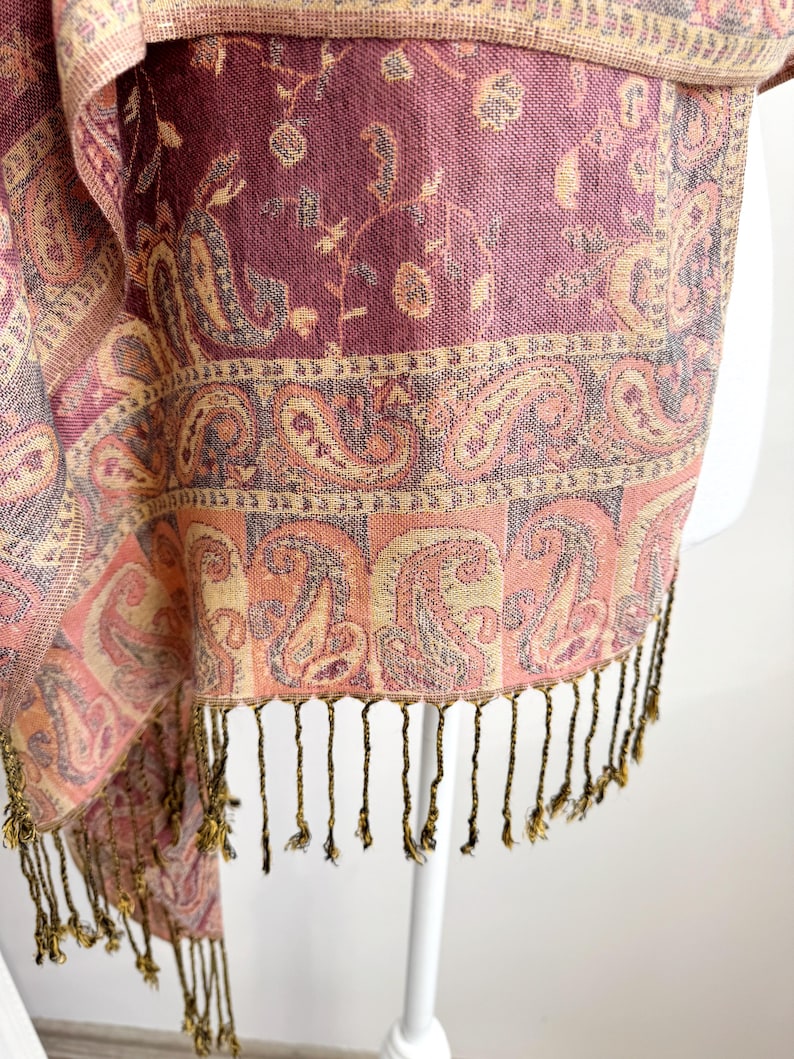 May include: A close-up of a paisley patterned scarf in shades of pink, peach, and purple. The scarf has a fringed edge with gold-colored tassels. The fabric has a woven texture and a decorative border.