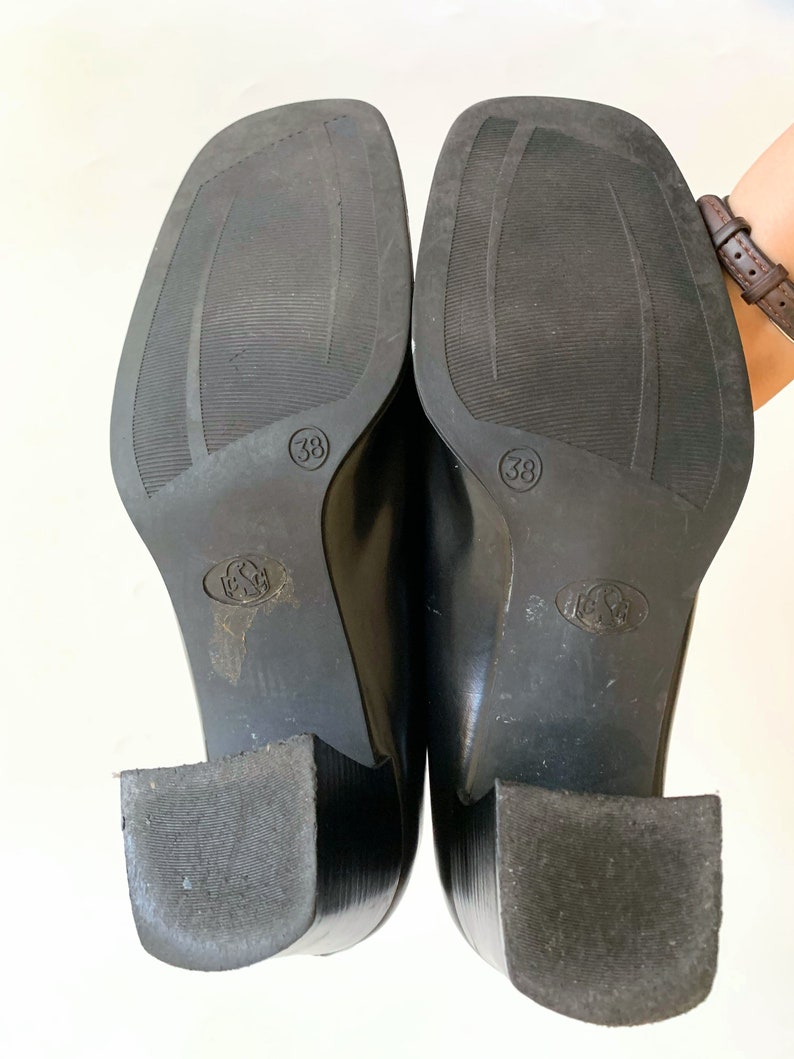 May include: A pair of black leather shoes with square toes and block heels. The soles are visible, showing size 38 and a textured pattern. The heels are approximately 2 inches high.