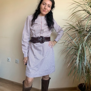 Puede incluir: Un vestido a rayas con un cinturón de cuero marrón y una hebilla grande. El vestido tiene mangas largas y un cuello. La persona lleva botas marrones.
