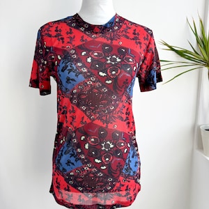 Puede incluir: Top de manga corta con estampado rojo, azul y burdeos. La tela transparente presenta un diseño floral y geométrico repetitivo. El top tiene un cuello alto y se muestra en un maniquí.