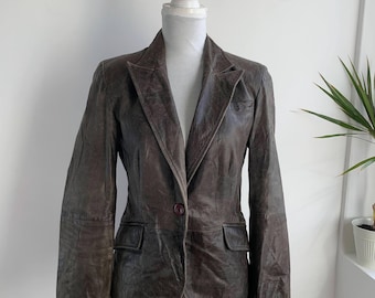 May include: A brown leather blazer with a single-button closure and two front pockets. The jacket is displayed on a mannequin against a white background. The blazer has a classic lapel and a tailored fit.