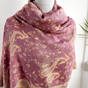 May include: A burgundy shawl with a paisley pattern in beige, gray, and light pink. The shawl is draped over a white form, showcasing the intricate floral and curved designs. The edges have a fringe.