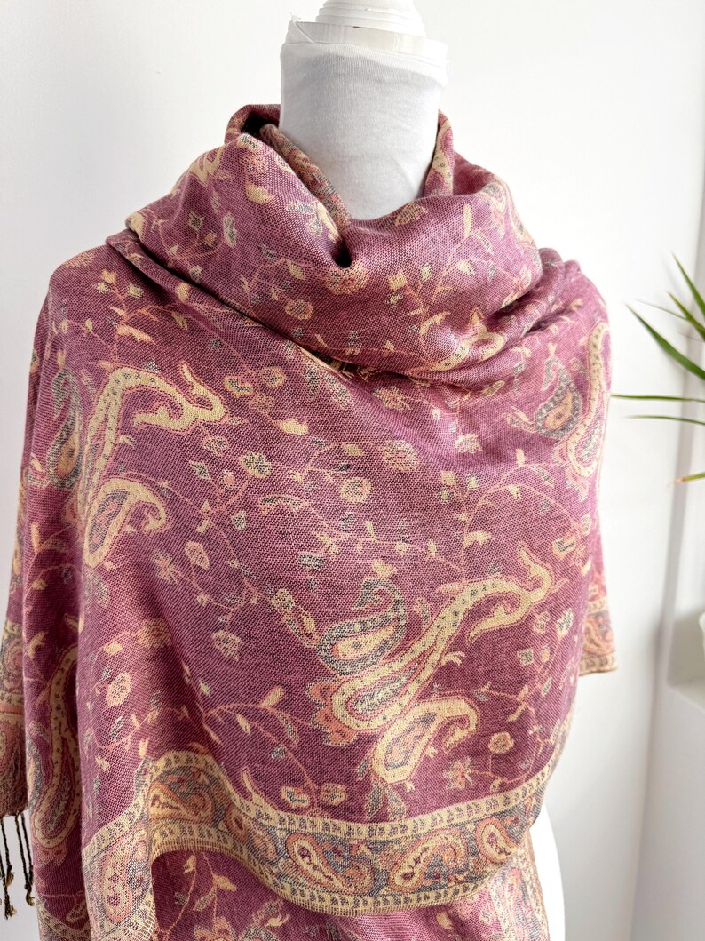 May include: A burgundy shawl with a paisley pattern in beige, gray, and light pink. The shawl is draped over a white form, showcasing the intricate floral and curved designs. The edges have a fringe.