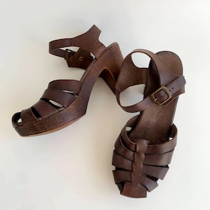 Könnte beinhalten: Ein Paar braune Ledersandalen mit Absatz und geflochtenem Design. Die Sandalen haben einen Schnallenverschluss und eine Holzplattform.