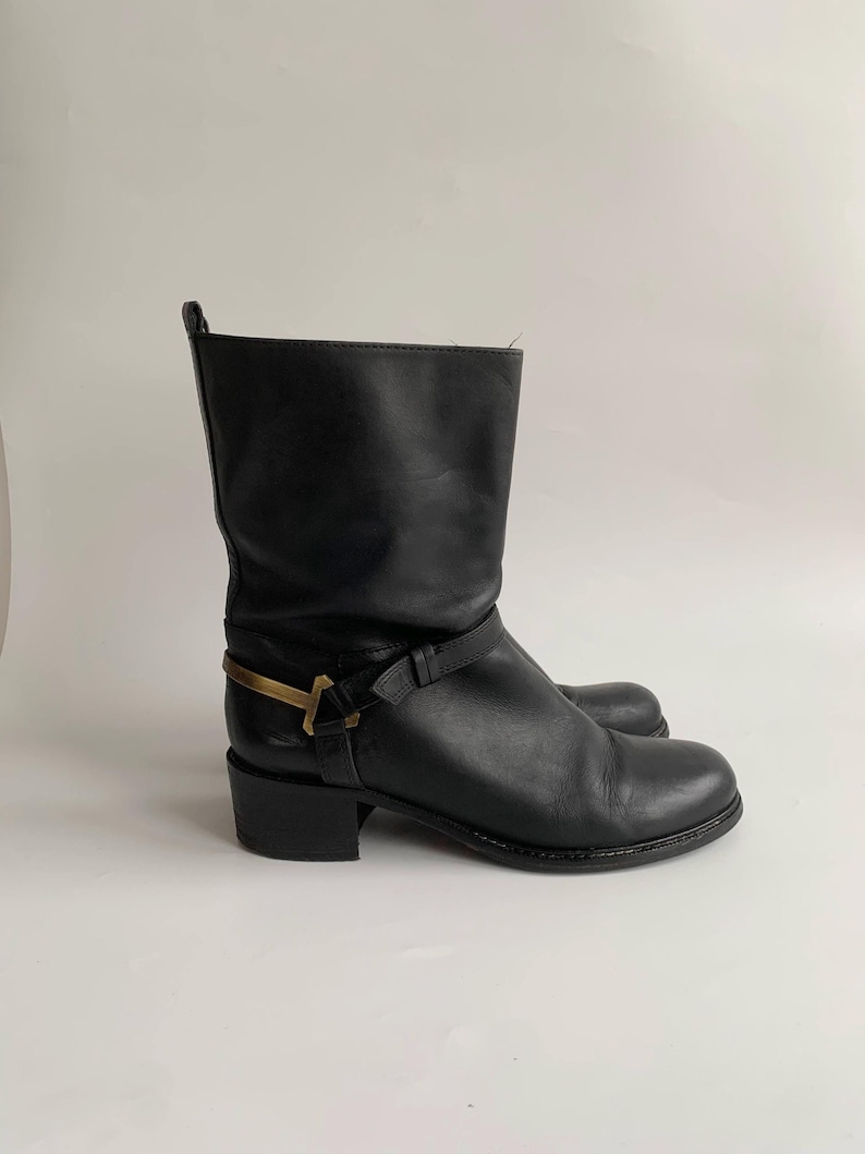 May include: A black leather mid-calf boot featuring a rounded toe, a low block heel, and a decorative strap across the ankle secured with a metallic gold-toned hardware accent. The boot has a pull tab at the top rear and a smooth finish.