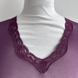 May include: A sheer, plum-colored top with a V-neckline. The neckline is trimmed with a delicate, dark purple lace. The top is draped over a white turtleneck, creating a contrast in color and texture. The fabric appears lightweight.