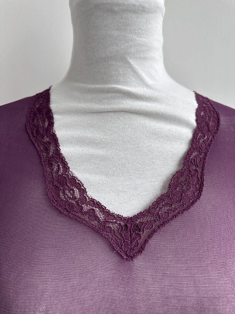 May include: A sheer, plum-colored top with a V-neckline. The neckline is trimmed with a delicate, dark purple lace. The top is draped over a white turtleneck, creating a contrast in color and texture. The fabric appears lightweight.