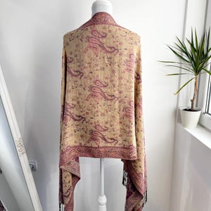May include: A beige shawl with a paisley pattern in shades of pink and burgundy is draped over a white mannequin. The shawl has a burgundy border and tassels at the ends. The background is a plain white wall.