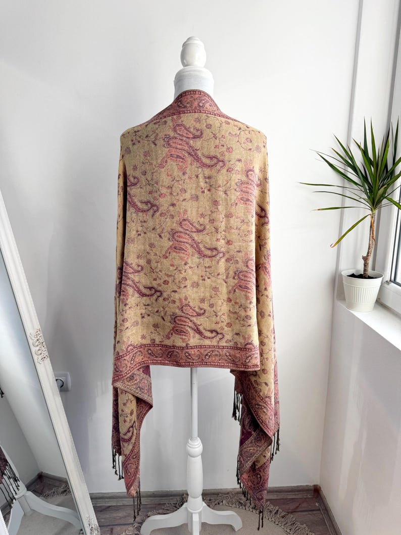 May include: A beige shawl with a paisley pattern in shades of pink and burgundy is draped over a white mannequin. The shawl has a burgundy border and tassels at the ends. The background is a plain white wall.