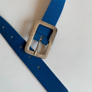 Pu&ograve; includere: Una cintura in pelle blu con una fibbia argentata. La cintura &egrave; piegata a met&agrave;, con la fibbia al centro dell'immagine.