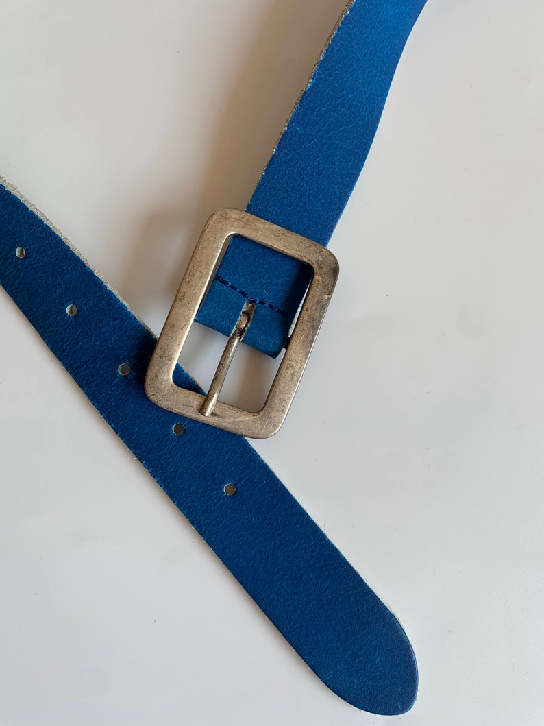 Pu&ograve; includere: Una cintura in pelle blu con una fibbia argentata. La cintura &egrave; piegata a met&agrave;, con la fibbia al centro dell'immagine.