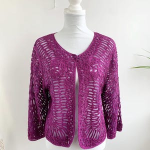 May include: A deep purple, open-front cardigan with a delicate, open-weave crochet design. The three-quarter sleeves and the intricate floral and geometric patterns add a touch of elegance. The cardigan is displayed on a mannequin.