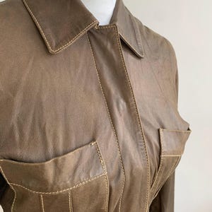 Op de afbeelding: Bruine leren jas met twee voorzakken en een kraag. De jas heeft zichtbare stiksels in een lichtere bruine kleur, wat een contrasterend detail toevoegt. De jas wordt gepresenteerd op een witte etalagepop.