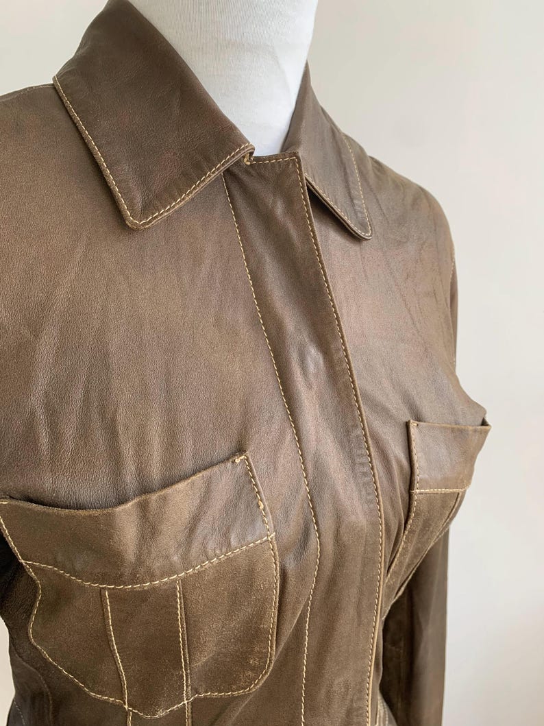 Op de afbeelding: Bruine leren jas met twee voorzakken en een kraag. De jas heeft zichtbare stiksels in een lichtere bruine kleur, wat een contrasterend detail toevoegt. De jas wordt gepresenteerd op een witte etalagepop.