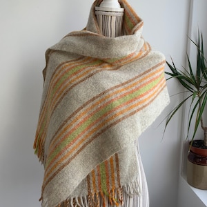 May include: A gray and white striped scarf with orange and green accents. The scarf has a fringe border and is draped over a mannequin.