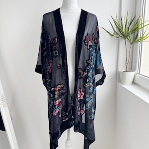 May include: A sheer black kimono-style jacket with a floral design in pink, blue, and green. The jacket has black velvet trim and is displayed on a white mannequin. The garment is lightweight and appears to be made of a delicate fabric.