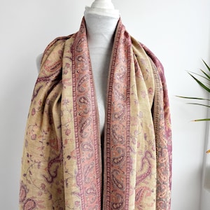 May include: A beige and pink paisley patterned scarf draped over a mannequin. The scarf features a central pink border with a paisley design, and a beige background with floral and paisley motifs. The scarf is made of a soft, flowing fabric.