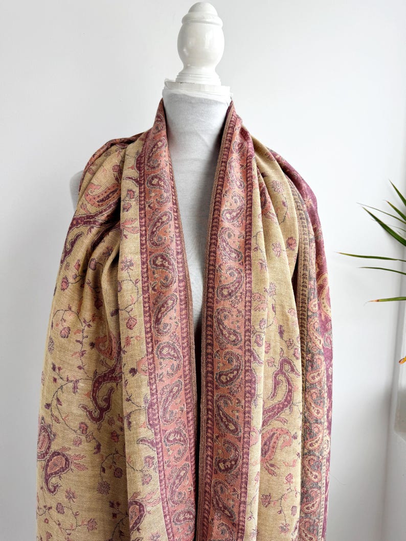 May include: A beige and pink paisley patterned scarf draped over a mannequin. The scarf features a central pink border with a paisley design, and a beige background with floral and paisley motifs. The scarf is made of a soft, flowing fabric.