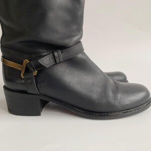 May include: A black leather boot featuring a low block heel, a rounded toe, and a decorative black leather strap wrapped around the ankle. The strap is secured with a metallic gold-toned hardware accent shaped like a T on the outer side.