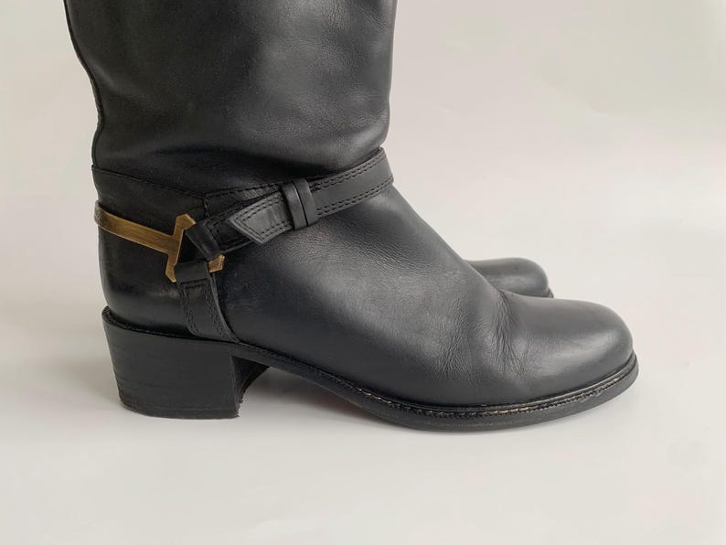 May include: A black leather boot featuring a low block heel, a rounded toe, and a decorative black leather strap wrapped around the ankle. The strap is secured with a metallic gold-toned hardware accent shaped like a T on the outer side.