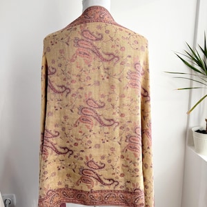 May include: A beige shawl with a paisley pattern in shades of pink and burgundy. The shawl is draped over a mannequin, showcasing the intricate floral and paisley design. The edges are finished with a decorative border and tassels.