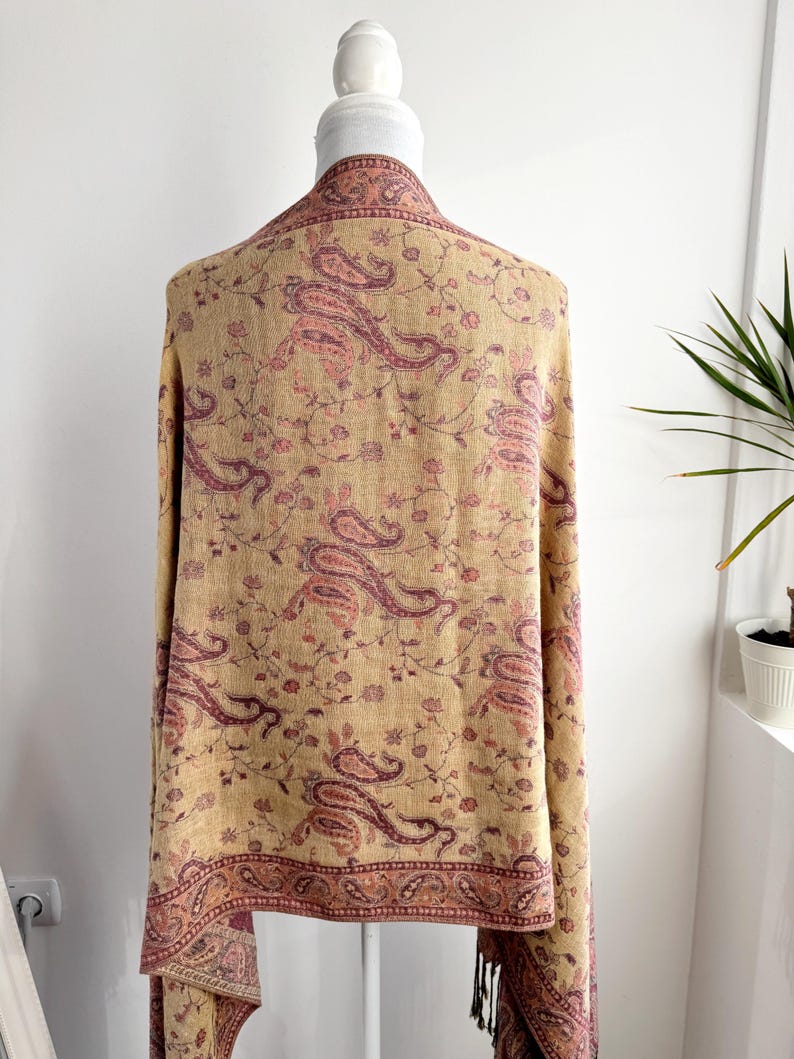 May include: A beige shawl with a paisley pattern in shades of pink and burgundy. The shawl is draped over a mannequin, showcasing the intricate floral and paisley design. The edges are finished with a decorative border and tassels.