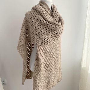 May include: A beige crochet shawl draped over a white mannequin. The shawl features a square pattern and a draped cowl neck. The open weave design is visible, and the shawl is displayed against a white background.