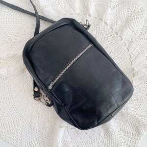 Pu&ograve; includere: Una borsa a tracolla in pelle nera con una cerniera argentata e una fibbia argentata. La borsa &egrave; su una superficie di uncinetto bianca.