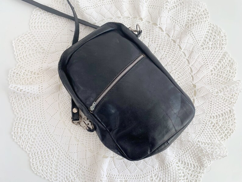 Pu&ograve; includere: Una borsa a tracolla in pelle nera con una cerniera argentata e una fibbia argentata. La borsa &egrave; su una superficie di uncinetto bianca.