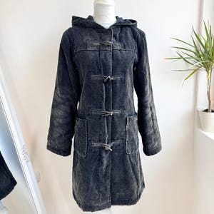 Op de afbeelding: Een donkergrijze corduroy jas met een capuchon en knevelsluitingen. De jas heeft twee voorzakken en wordt op een mannequin getoond. De jas is een lange lengte en heeft lange mouwen.