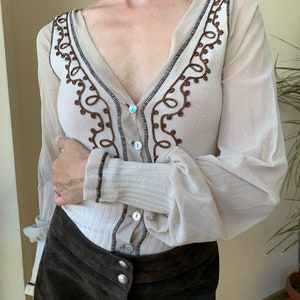 May include: A beige sheer long-sleeve button-up shirt with brown embroidered detailing around the neckline and cuffs. The shirt is paired with a brown suede skirt with button closures.