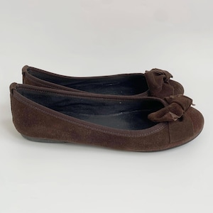 May include: A pair of brown suede ballet flats with a bow detail on the toe. The shoes have a rounded toe and a flat sole. The flats are a solid brown color and appear to be unworn.