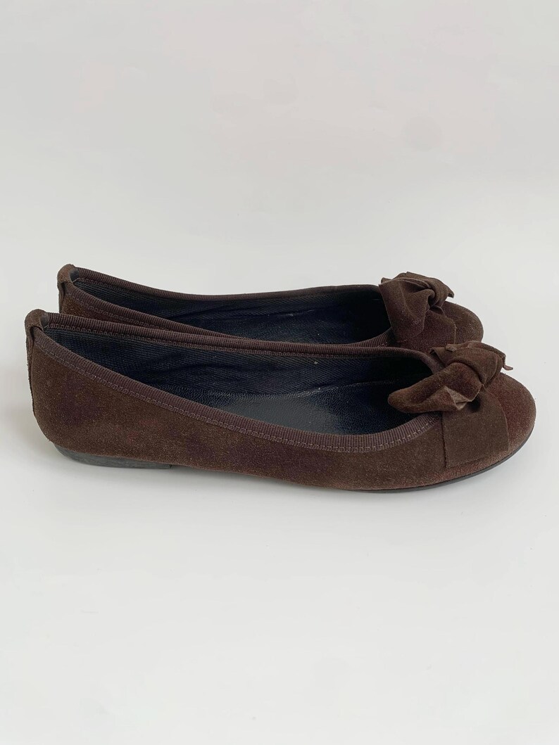 May include: A pair of brown suede ballet flats with a bow detail on the toe. The shoes have a rounded toe and a flat sole. The flats are a solid brown color and appear to be unworn.