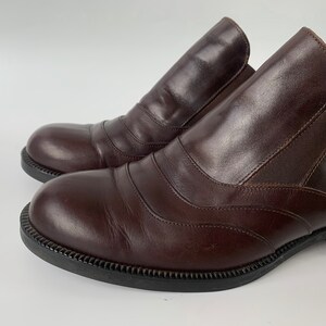 K&ouml;nnte beinhalten: Ein Paar braune Slipper aus Leder mit einem dekorativen Steppdetail. Die Schuhe haben eine abgerundete Spitze und einen niedrigen Absatz.