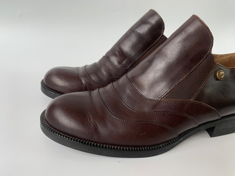 K&ouml;nnte beinhalten: Ein Paar braune Slipper aus Leder mit einem dekorativen Steppdetail. Die Schuhe haben eine abgerundete Spitze und einen niedrigen Absatz.