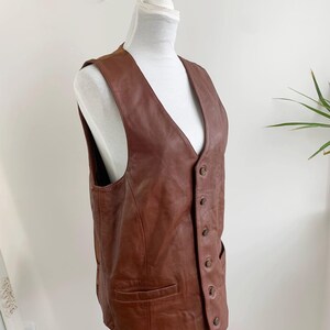May include: A brown leather vest with a V-neck and button closure. The vest has two front pockets and is displayed on a white mannequin. The leather appears to be well-worn, with a rich, warm tone. The vest is a classic piece of apparel.