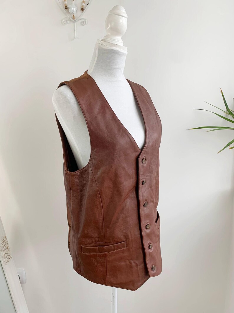 May include: A brown leather vest with a V-neck and button closure. The vest has two front pockets and is displayed on a white mannequin. The leather appears to be well-worn, with a rich, warm tone. The vest is a classic piece of apparel.