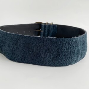 May include: A wide, navy blue belt with a textured finish. It features a metal buckle and adjustment holes. The belt is probably made of leather or a similar material.